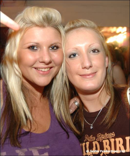 Two smiling young women with long blonde hair, one in a purple top and one in a brown printed T-shirt