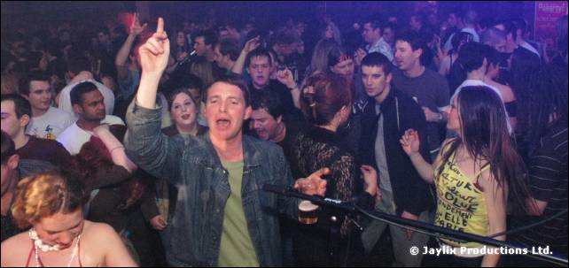 Packed crowd on a dance floor, with a man in a denim jacket raising one arm