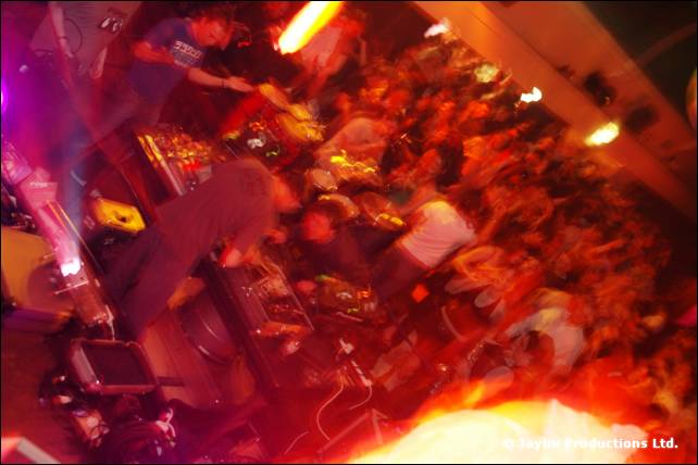 Blurred, red-lit view over a crowd towards DJ decks, with bright flame effects at the top