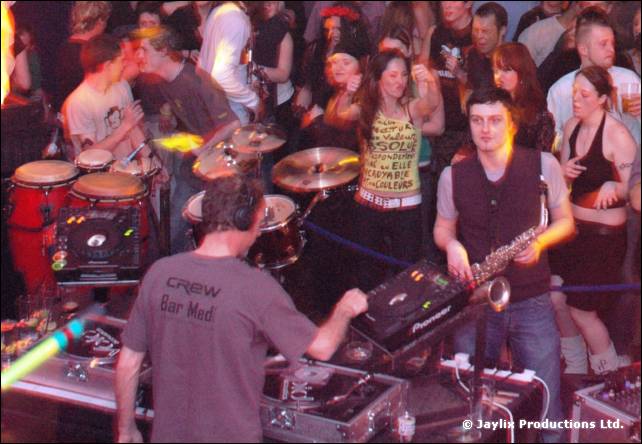 DJ in a Crew Bar Med T-shirt at the decks, with a saxophonist, drum kit and dancing crowd behind