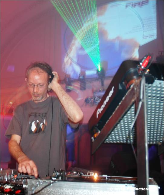 DJ in a grey FIRE T-shirt and headphones at a Pioneer mixer, with a green laser and projection behind