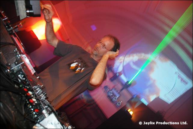 DJ in a grey T-shirt and headphones reaches up at the decks under a green laser beam