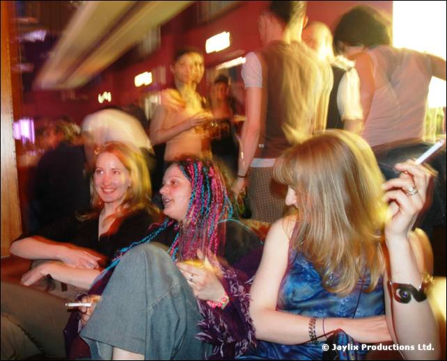 Three seated women on a sofa, one holding a cigarette, with people standing and blurred lights behind