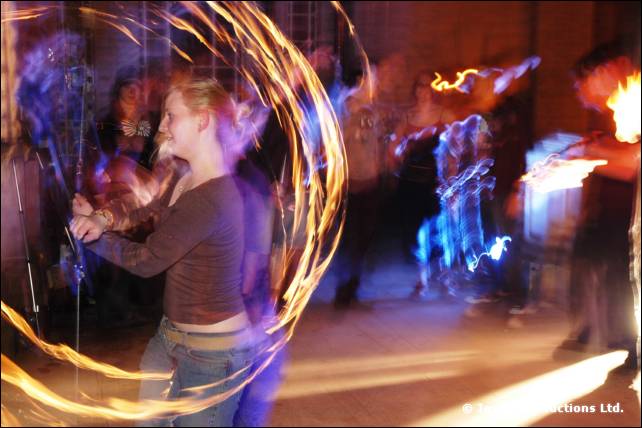 Person spins a ring of fire poi in a dim hall, with blurred onlookers behind