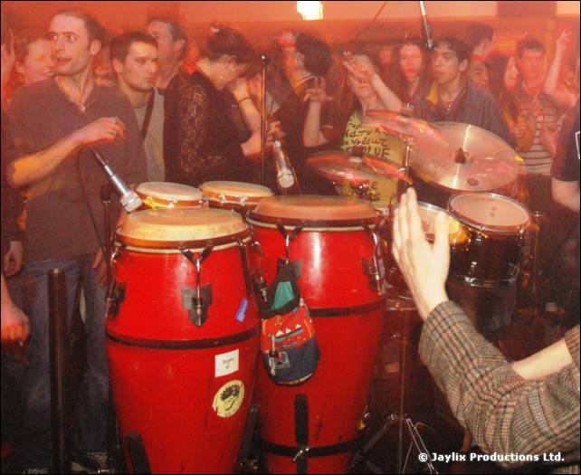 Red conga drums and a drum kit in a packed room, people crowded behind and a hand raised in the foreground