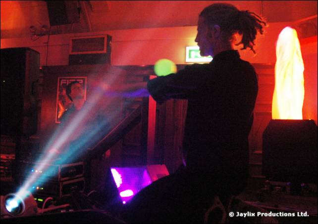Silhouetted person with dreadlocks holds a glowing green ball on a stage lit by beams of light