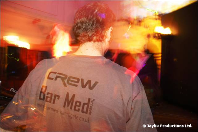 Back of a person in a grey Crew Bar Med T-shirt, with orange flame blur beyond