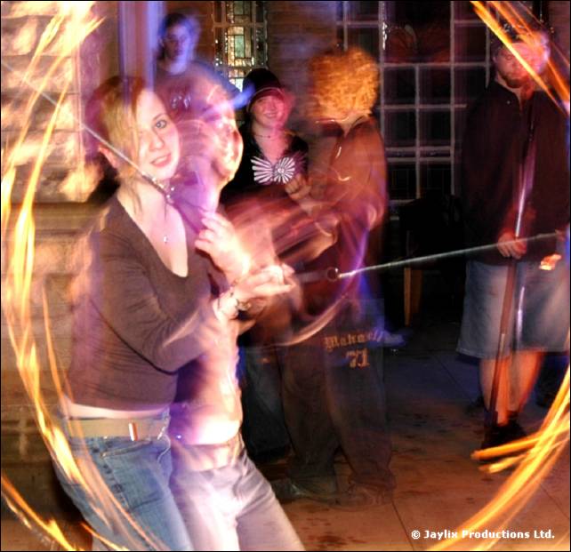 Woman in a brown top dances, with a blurred fire staff trail circling her and people watching behind