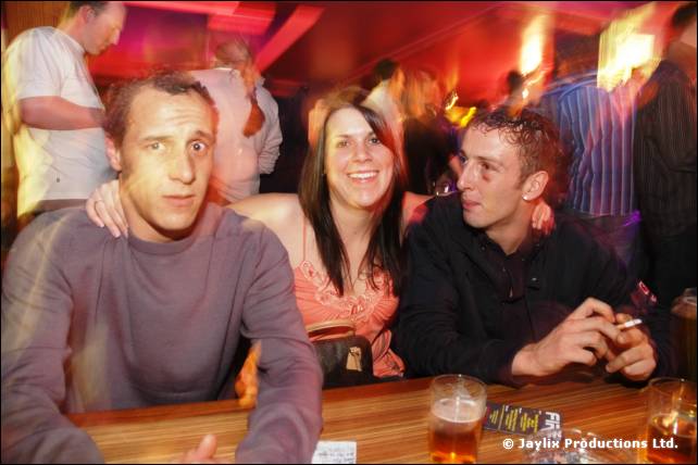 Three people sit at a wooden bar with drinks, the man on the right holding a cigarette