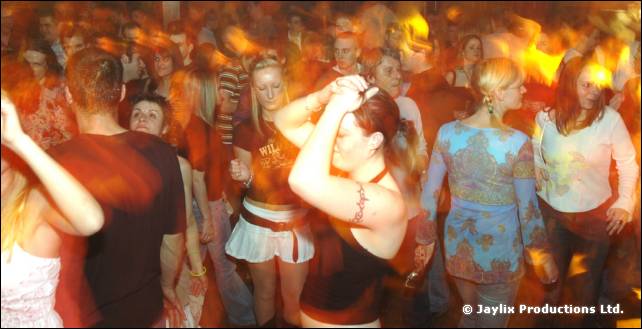 Crowd dancing in a busy hall, with a woman in a white skirt and black top in the foreground