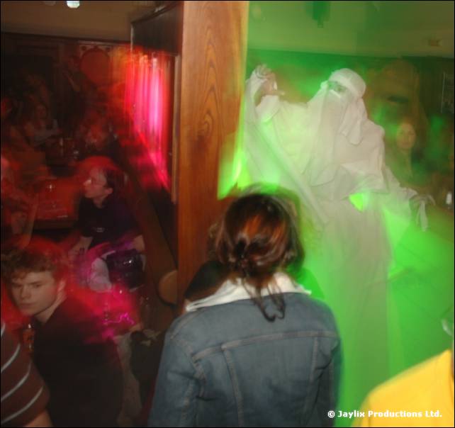 Person in a white robe and veil stands lit by green light above a dark crowd, with a woman in a denim jacket in front