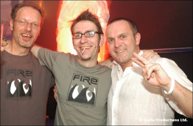 Three men stand together smiling, two in grey FIRE T-shirts and one in a white shirt making a peace sign