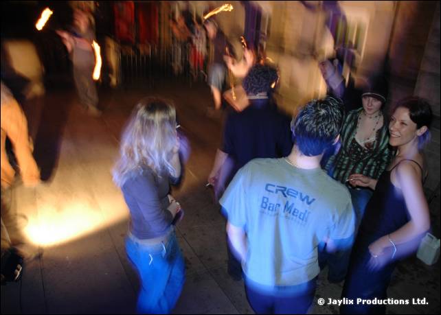 View from behind of dancers on a floor, one in a Crew Bar Med T-shirt, in dim blurred light