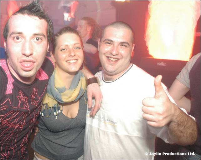 Three smiling people pose together, one making a thumbs up, in front of an orange lit column