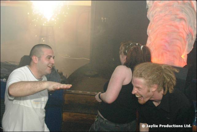 A man in a white T-shirt points as a woman and a man with dreadlocks laugh, in a smoky, orange lit room