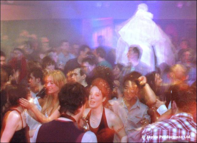 Crowd of dancers in purple light, with a tall figure in white robes above them at the back
