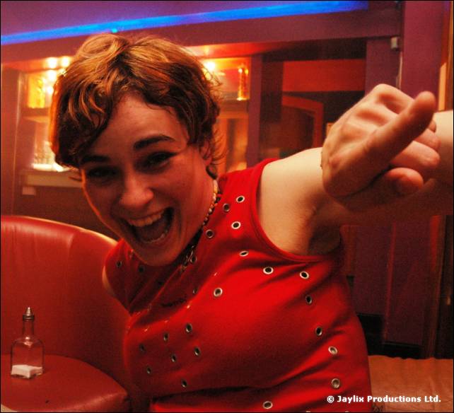 Woman in a red studded top laughs and points at the camera, seated by a red booth