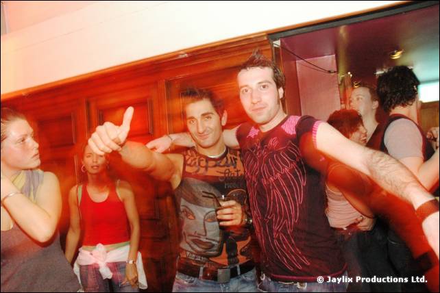 Two men pose with arms around each other, one giving a thumbs up, with people behind in a wood panelled room
