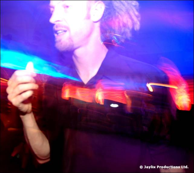 Blurred man with curly hair, lit in blue and purple, raises a hand