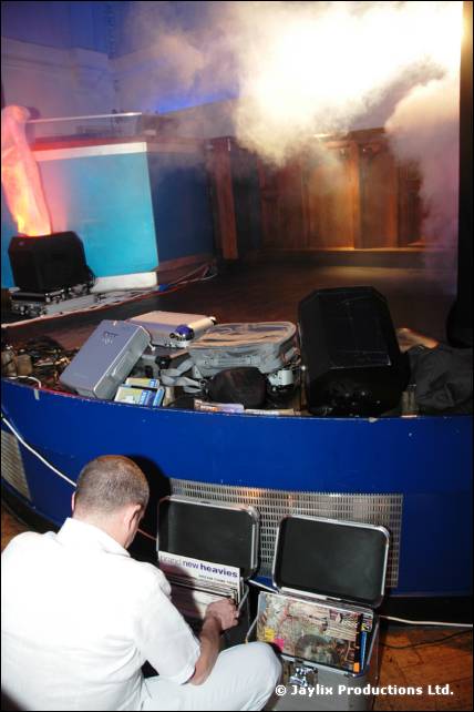 Man in white sits on the floor at open equipment cases in front of a blue curved bar, with smoke rising behind