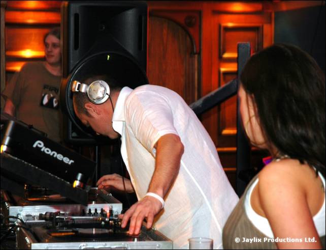 DJ in a white shirt and headphones at Pioneer decks, with a dark haired woman beside him