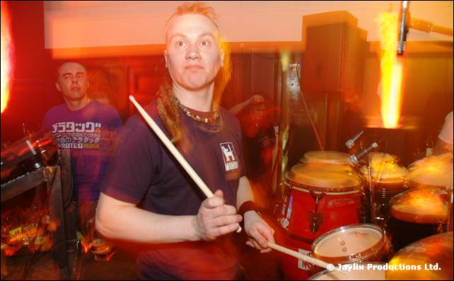 Drummer with a mohican holds two sticks behind a red drum kit, with a flame effect beside him