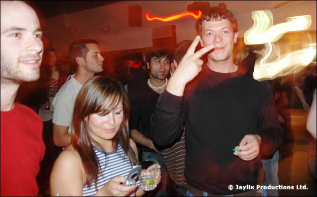 Group of people at a club night, one in a dark top making a peace sign, a woman looking at her phone