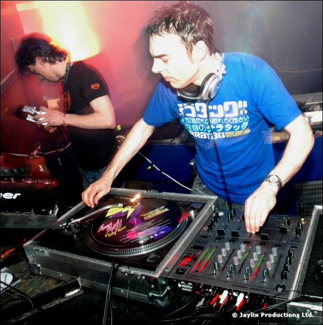 DJ in a blue T-shirt with Japanese print mixes at turntables, with another person beside him