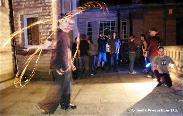 Person twirls fire poi on a stone terrace at night, with onlookers standing behind