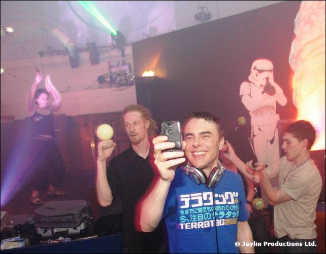DJ in a blue Japanese print T-shirt and headphones holds up a phone, with a Stormtrooper cutout behind