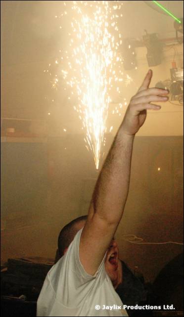 Raised arm points upwards beside a fountain of sparks, in a smoky room