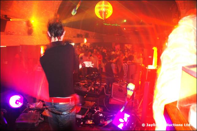 Person in black with their back to the camera stands at equipment on stage, facing a crowd under purple lights