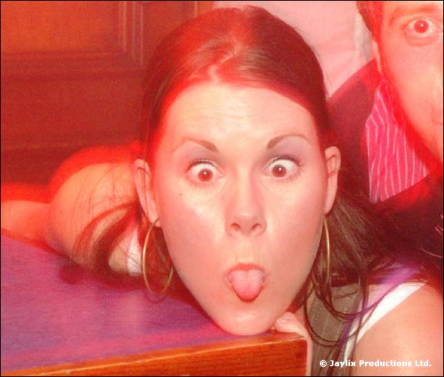 Woman pulls a face with wide eyes and tongue out, lying against a table, with a man's face behind her
