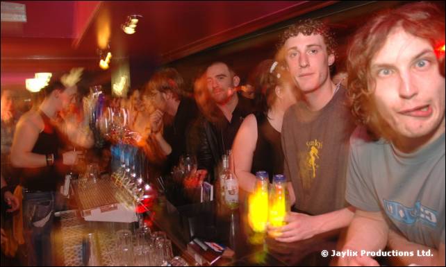 Crowded bar with two men in the foreground, one pulling a face, and glasses of yellow drinks on the bar