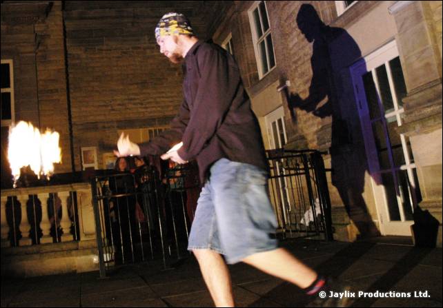 Person in a bandana and denim shorts walks along a stone terrace with a flaming torch, casting a shadow