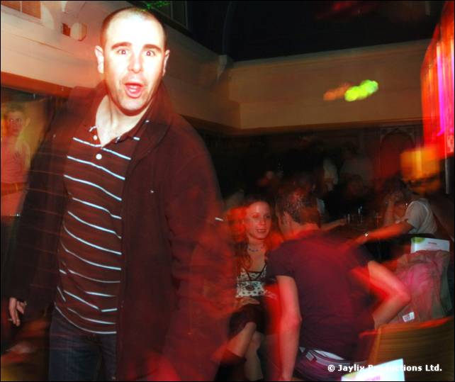 Man in a striped top and dark jacket looks towards the camera, with blurred dancers behind
