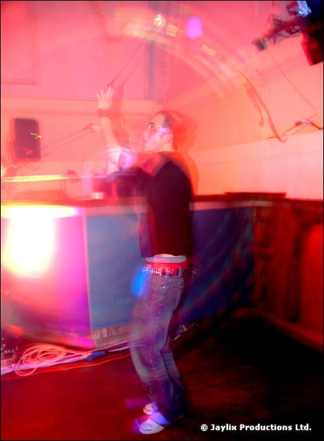 Blurred person in dark clothes stands beside a lit DJ booth in a hall with a pink glow