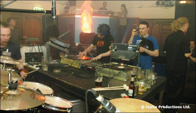 DJs and drummers at a booth with drum kit cymbals in front and a flame effect behind