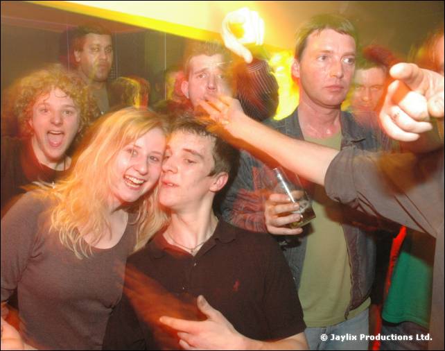 Group of people laughing and leaning together in a crowd, with one hand pointing at the camera