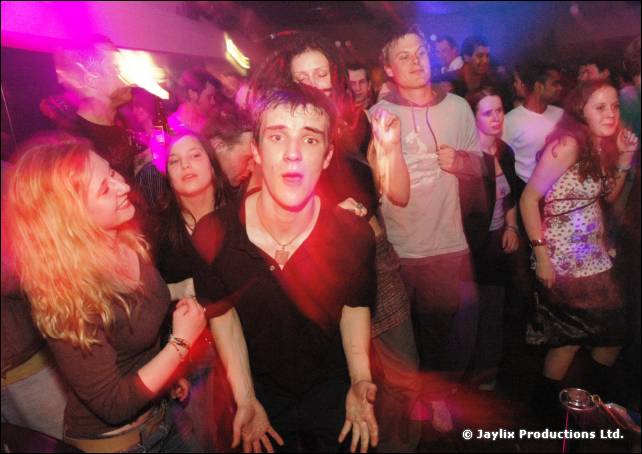 Crowd on a dance floor in purple light, with a young man in black looking at the camera