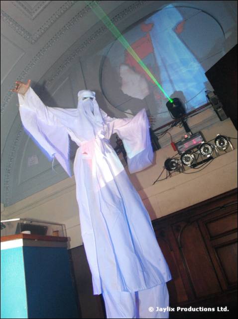 Stilt walker in a white robe and veil raises one arm under a green laser, near DJ equipment