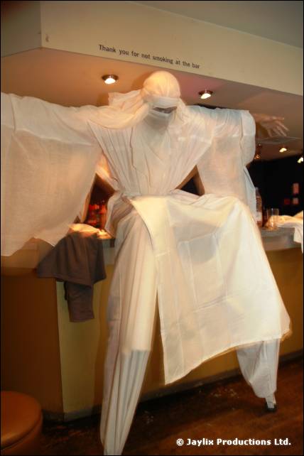 Stilt walker in a white robe and veil stands with arms out beside a bar