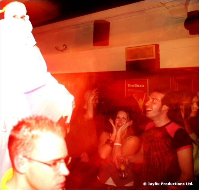 Stilt walker in white at the left, above a seated crowd, with a man in glasses in the foreground