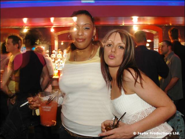 Two women pose with cocktails, one in a white top and one in a white lace top, in a bar