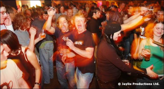 Crowd dancing with raised arms and drinks, with three men smiling at the front