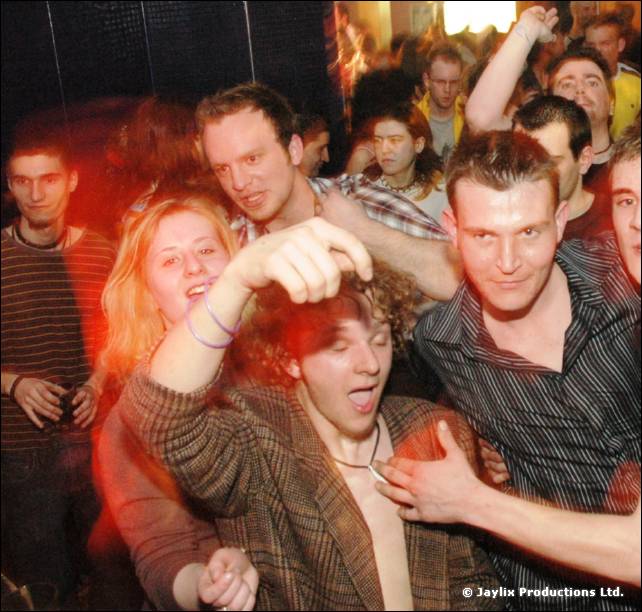 Crowded group of people, one man with open shirt and curly hair in the foreground, with a woman behind him