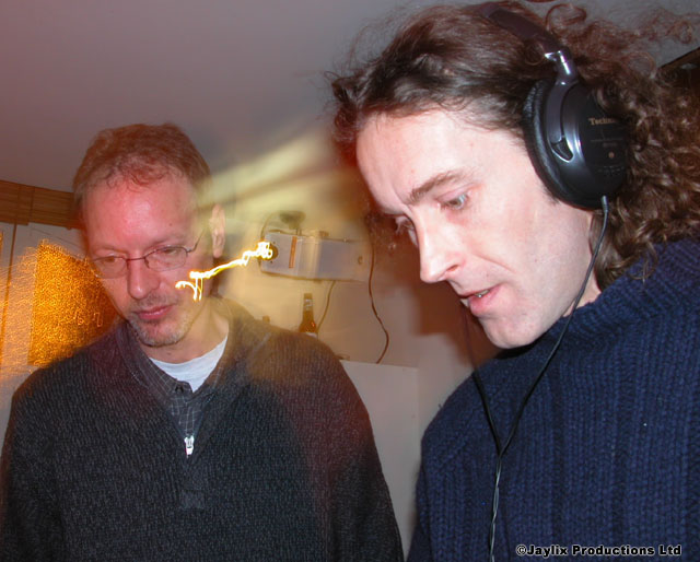 Two men look down at something, the one on the right wearing headphones