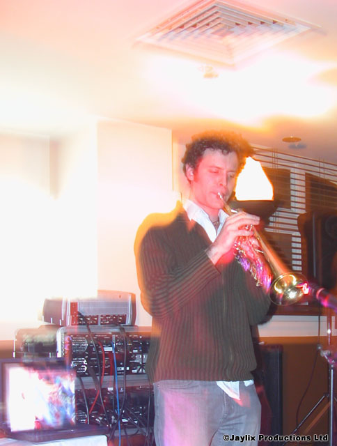 A man plays a trumpet in front of audio equipment in an overexposed room