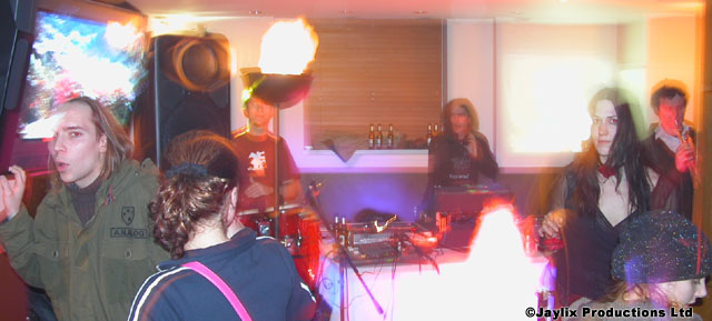 People gather near a DJ booth with flaming torches and a drum kit in a bar