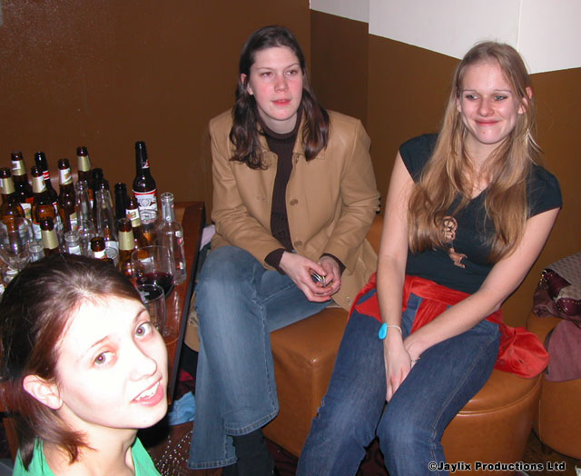 Three women sit together on a leather bench beside a table of bottles